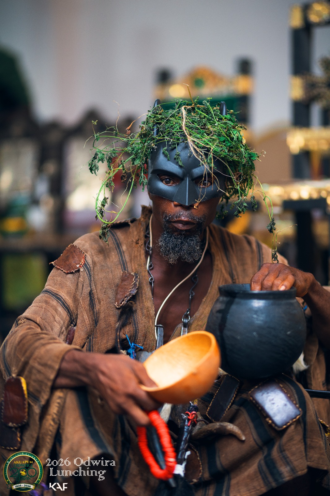 A masked custodian crowned with fresh herbs holds a black pot and a calabash