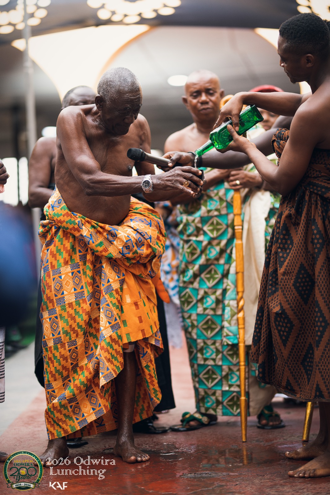 An elder pours libation from a bottle held by an attendant, surrounded by chiefs in kente