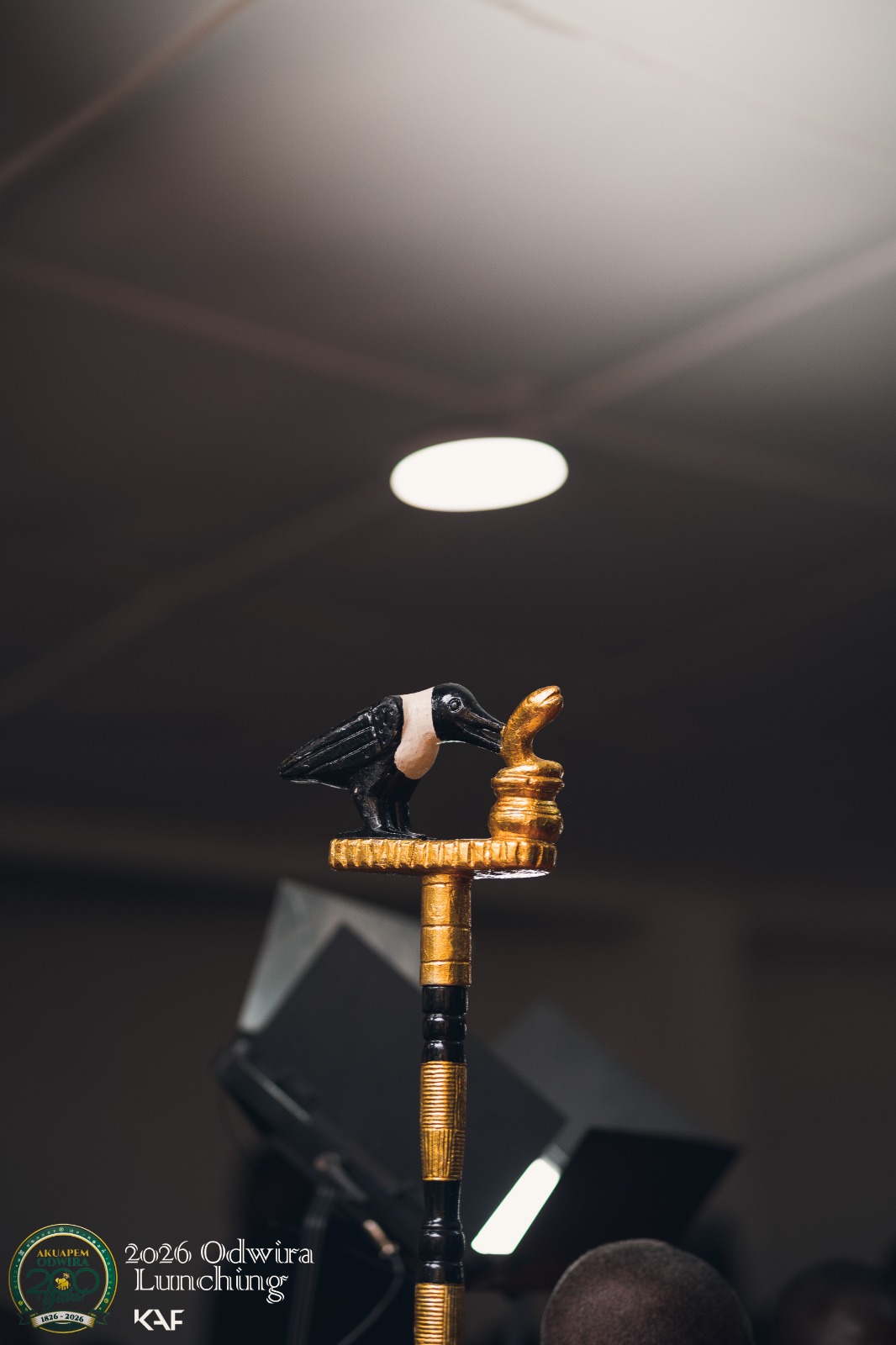 A black-and-white crow finial facing a gold form, on a linguist staff under a spotlight